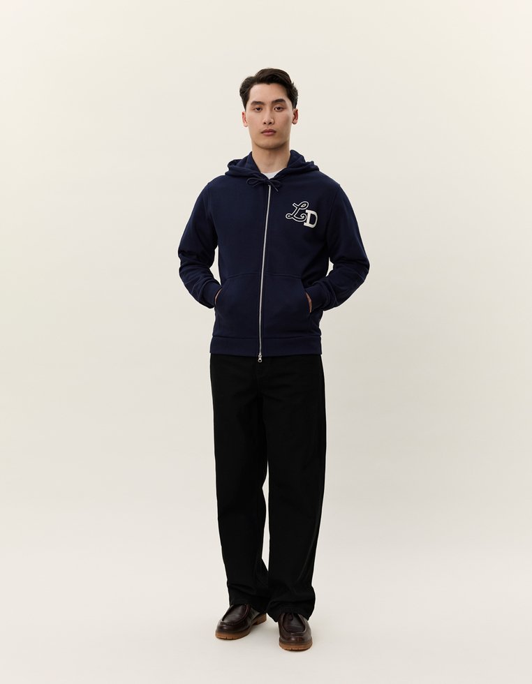 Felpa con zip di colore blu navy e coulisse, logo "L&D" bianco sul petto, abbinata a pantaloni neri e scarpe marroni. Tessuto dalla texture liscia.