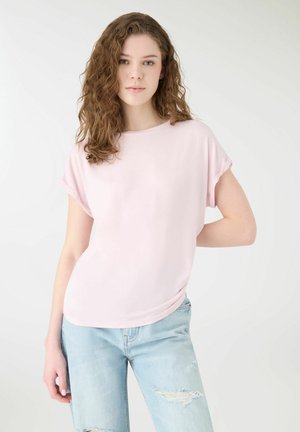 Młoda kobieta o kręconych włosach nosi bladoróżowy krótki rękaw i jasnoniebieskie podarte dżinsy, stoi na tle jednolitego białego tła.