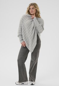 Hellgrauer gestrickter Poncho mit einem offenen Webmuster, der einen weiten Ausschnitt und lange Ärmel hat, kombiniert mit dunkelgrauen Schlaghosen und Sneakers.