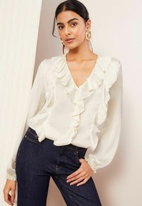 Weiße Bluse mit gerafftem Ausschnitt und langen Ärmeln, kombiniert mit dunkelblauen Jeans. Die Bluse hat einen weichen, strukturierten Stoff und zarte Manschetten.