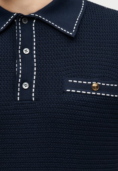 Σκούρο μπλε πλεκτό πόλο μπλουζάκι με λευκή διακόσμηση στον γιακά, την πατιλέτα και το καπάκι της τσέπης, με μαύρα και χρυσά κουμπιά.