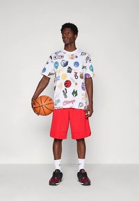 Balta grafiska T-krekls ar krāsainiem sporta logotipiem, saskaņota ar sarkaniem basketbola šortiem. Modelis tur rokās basketbola bumbu un valkā melnas sporta apavus.