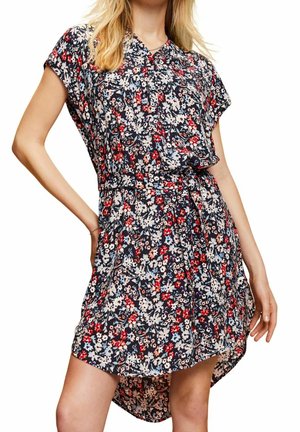 Femme portant une robe à manches courtes à motifs floraux avec des fleurs rouges, blanches et bleues, un décolleté en V et un ourlet légèrement asymétrique.