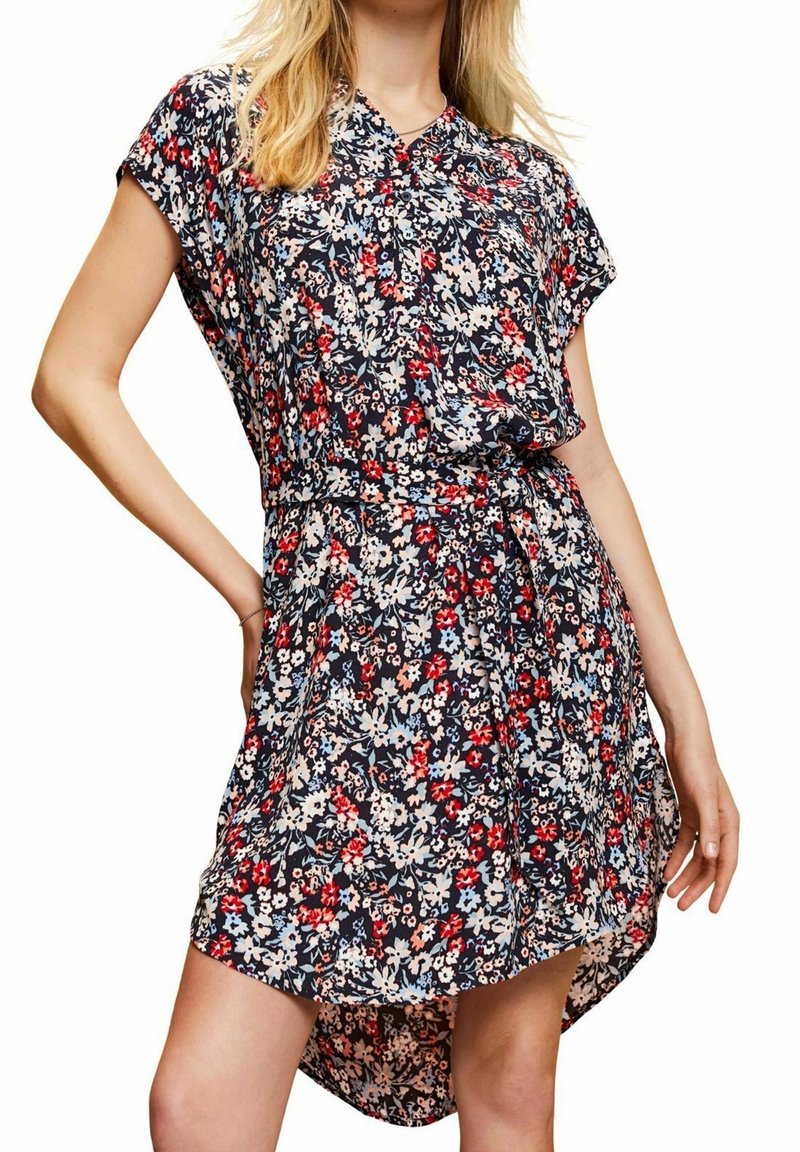 Femme portant une robe à manches courtes à motifs floraux avec des fleurs rouges, blanches et bleues, un décolleté en V et un ourlet légèrement asymétrique.