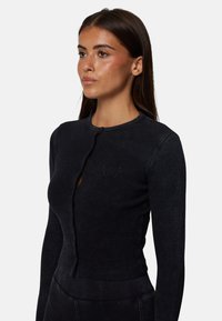 Czarna, żeberkowana bluzka z długim rękawem, z okrągłym dekoltem, z lekkim detalem w kształcie dziurki oraz dyskretną haftowaną logo na piersi.