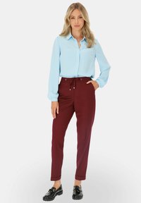 Kobieta w jasnoniebieskiej bluzce, burgundowych spodniach z troczkiem i czarnych loafersach, stojąca z jedną ręką w kieszeni na białym tle.