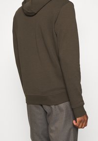Hoodie verde oliva con cappuccio regolabile e polsini a coste, realizzato in tessuto morbido. Indossato sopra pantaloni a motivo quadri. Design semplice e aderente.