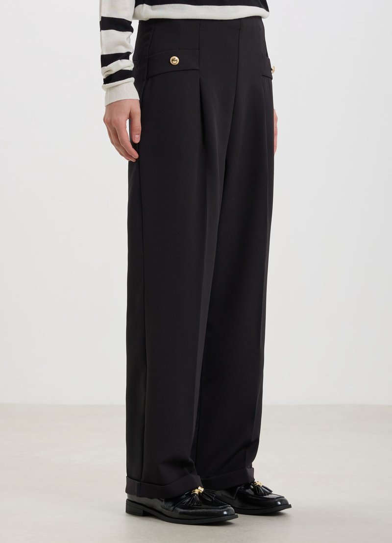 Pantalons larges noirs avec plis avant, deux poches latérales agrémentées de boutons dorés, associés à des mocassins noirs avec des détails à franges.
