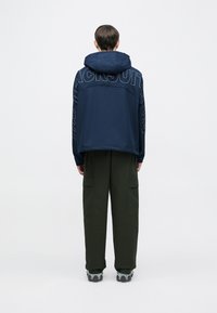 Tengeralattjáró kék kapucnis pulóver nagy grafikával a hátoldalán, olívazöld dzseki nadrággal és szürke tornacipővel párosítva. Hétköznapi, laza fazon.