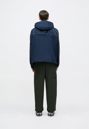 Sweatshirt azul-marinho com capuz e grande texto gráfico nas costas, combinado com calças cargo verde-oliva e sapatilhas cinzentas. Ajuste casual e descontraído.