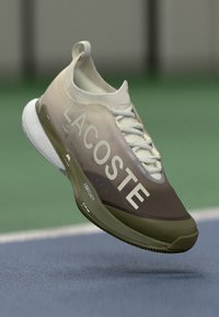 Sneakers Lacoste v béžové a hnědé barvě, s texturovaným svrškem, bílými tkaničkami a výrazným logem. Gumová podrážka se štíhlým designem.