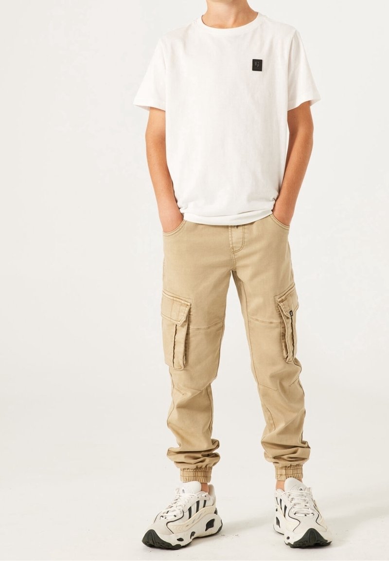 Personne portant une chemise blanche à manches courtes avec un petit logo, un pantalon cargo beige avec des poches, et des baskets blanches avec des motifs noirs, les mains dans les poches.