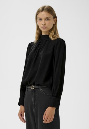 Blouse noire à manches longues avec un col montant, un design plissé et des boutons aux poignets, associée à un jean en denim foncé et une ceinture.