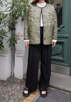 Personne portant une veste matelassée vert olive, un pull blanc, un pantalon large noir et des sandales plates-formes noires, debout à l'extérieur près d'une porte verte et de plantes murales.
