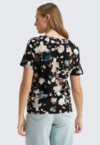 Donna con capelli lunghi che indossa una maglietta nera a maniche corte con un motivo astratto marrone, bianco e blu, abbinata a jeans azzurri chiari.