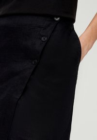 Jupe en lin noir avec fermeture à bouton sur le côté et une poche visible. Le tissu a un aspect légèrement texturé.