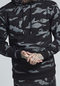 Black and gray camo pullover hoodie with a drawstring hood, ribbed cuffs, and hem; features a textured fabric and a subtle logo on the chest.