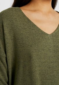 Maglione verde oliva a maglia con scollo a V, caratterizzato da una texture morbida e un motivo leggermente mélange. Nessun accessorio o dettaglio vistoso visibile.