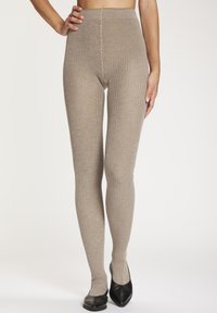 Nahaufnahme einer Person, die hoch taillierte, gerippte beige Strickleggings trägt, kombiniert mit schwarzen spitzen Ballerinas vor einfachem Hintergrund.