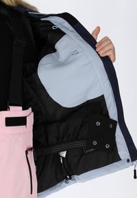 Różowe spodnie śniegowe z czarnym wykończeniem, wyposażone w kieszeń na zamek, regulowane paski i miękką niebieską podszewkę.