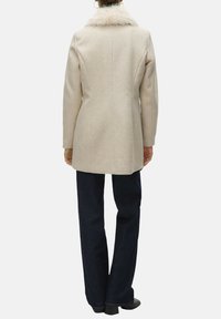 Manteau en laine beige avec col en fausse fourrure, coupe cintrée, coupe droite et manches longues, porté sur un jean bleu foncé à jambes larges.