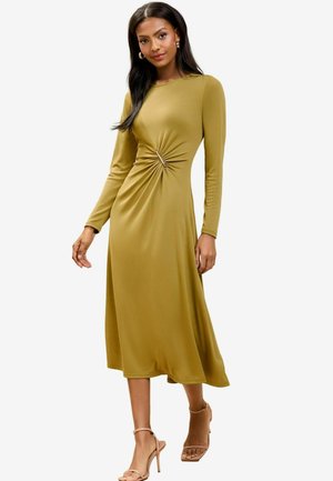 Rochie verde măslinie cu mâneci lungi și detaliu răsucit la talie, realizată dintr-un material neted. Asortată cu sandale cu toc nude, pentru un look complet.