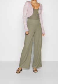 Roze cropped vest over een groene, getextureerde bodysuit en olijfgroene broek met wijde pijpen. Zwarte sandalen met hak maken de outfit compleet.