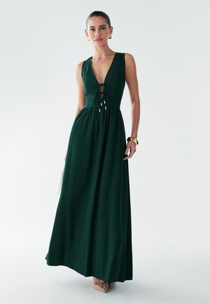 Maxi dress verde in tessuto morbido con scollatura a V profonda e vita arricciata, caratterizzata da una chiusura anteriore a fiocco e dettagli dorati. Completata da scarpe beige.