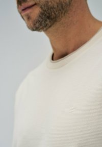 Suéter texturizado beige claro con cuello redondo y un sutil detalle de logo en el pecho. La vista cercana resalta el patrón de la tela y las costuras.