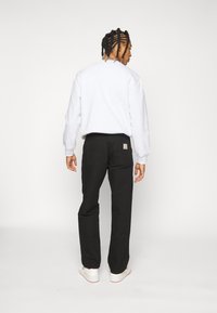 Grauer Sweatshirt mit gerippten Bündchen, kombiniert mit schwarzen Hosen, die über zwei Gesäßtaschen und ein Logo-Patch verfügen; Modell steht mit dem Rücken zur Kamera vor einem weißen Hintergrund.
