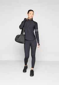 Black athletic long-sleeve top with zip collar and gray stripes, paired with black leggings and sneakers, with a black duffle bag.