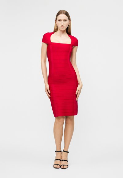 Žena stojící v přiléhavých červených šatech po kolena s krátkými rukávy a černých sandálech na vysokém podpatku se stříškami na jednoduchém pozadí.