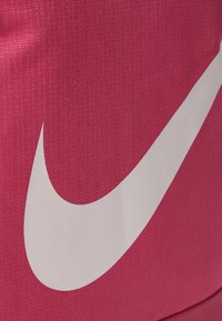 Roza tekstilija z teksturirano površino, ki prikazuje velik bel znak Nike swoosh. Oblikovanje poudarja preprostost in prepoznavnost blagovne znamke.