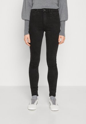 Jeans negros ajustados con una textura suave, cinturilla alta, bolsillos frontales y detalles de costura, combinados con zapatillas gris y un suéter gris.