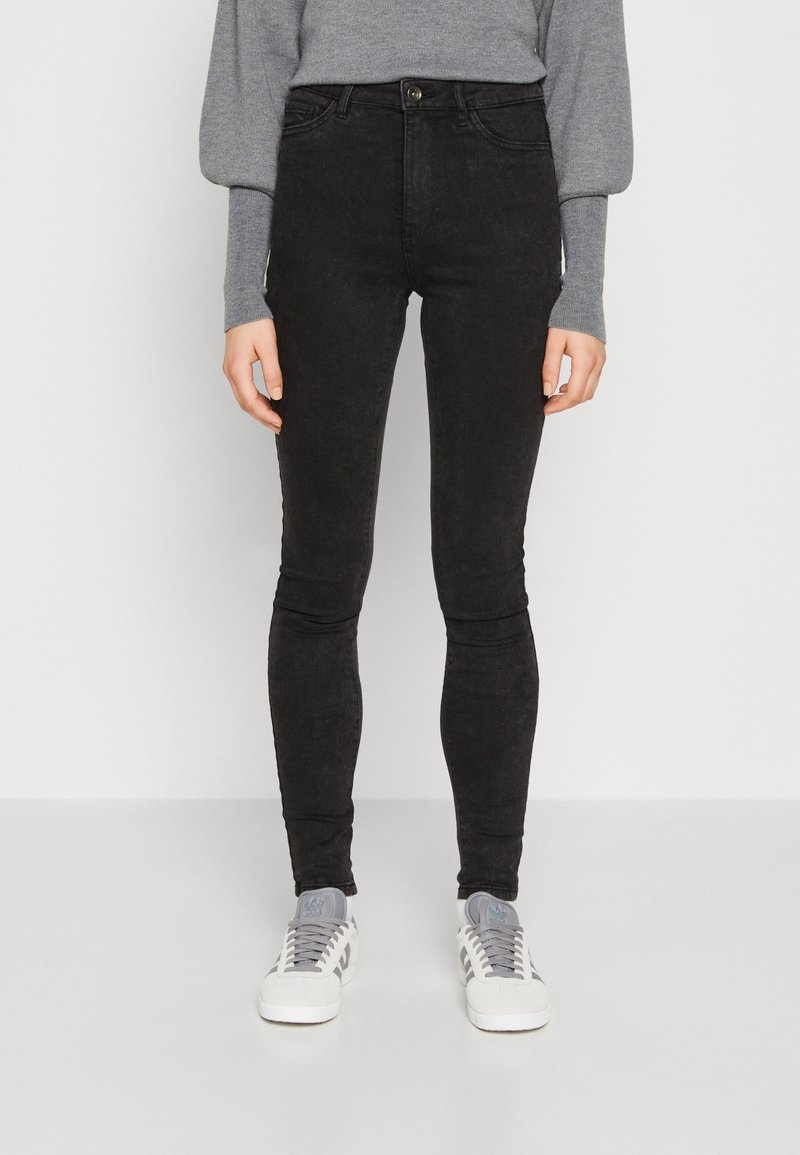 Jeans negros ajustados con una textura suave, cinturilla alta, bolsillos frontales y detalles de costura, combinados con zapatillas gris y un suéter gris.