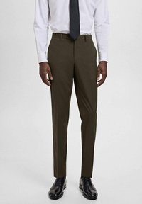 Man in een donkerbruine op maat gemaakte broek, witte overhemden, zwarte das en zwarte nette schoenen, staand tegen een effen achtergrond.