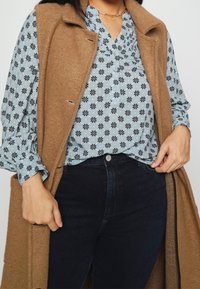 Gilet en laine tan porté sur un blouse à fleurs bleues avec des motifs noirs, associé à un jean foncé. Présente une fermeture à boutons et des manches longues.