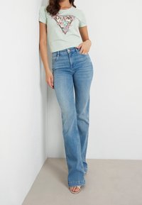 Světle zelené tričko s květinovým a flitrovým trojúhelníkovým vzorem, spárované s vysokými, rozšířenými světle modrými džíny a stříbrnými sandály.