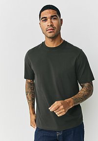 Tumši zaļš, īsām piedurknēm, kokvilnas T-krekls ar apaļu kaklu. Ar brīvu piegriezumu un gludu tekstūru. Valkāts kopā ar ziliem džinsiem.