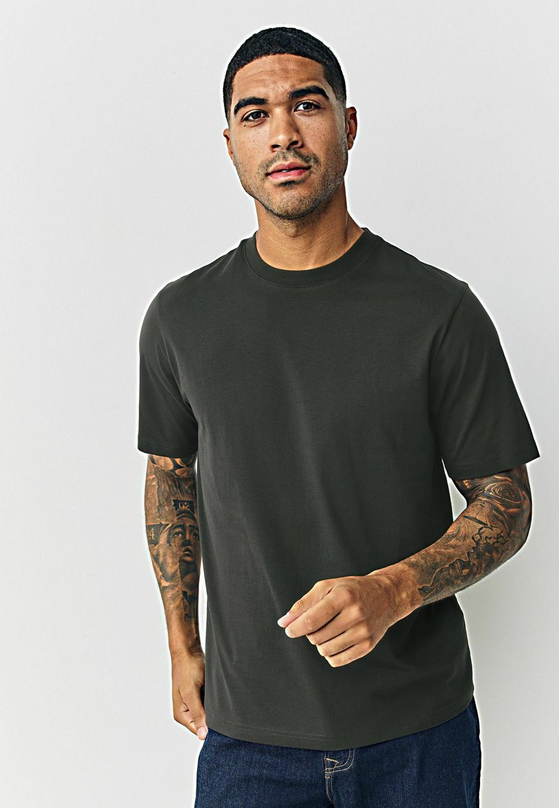 Tumši zaļš, īsām piedurknēm, kokvilnas T-krekls ar apaļu kaklu. Ar brīvu piegriezumu un gludu tekstūru. Valkāts kopā ar ziliem džinsiem.