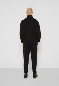 Černá mikina s vysokým límcem, volný střih a žebrované manžety, v kombinaci s černými teplákovými kalhotami a černou obuví.