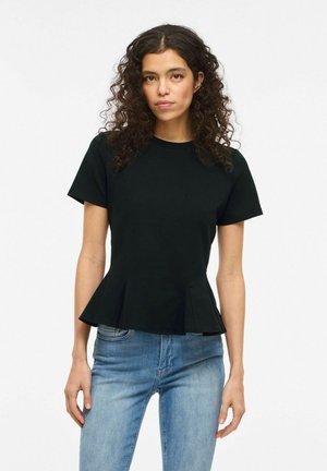 Femme aux cheveux bouclés portant un haut noir à manches courtes et basque, ainsi qu'un jean bleu clair, debout devant un fond blanc uni.