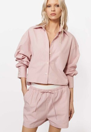 Kobieta ubrana w luźną różowo-białą koszulę w paski zapinaną na guziki oraz pasujące spodenki na sznurku, ręce w kieszeniach, neutralne tło.