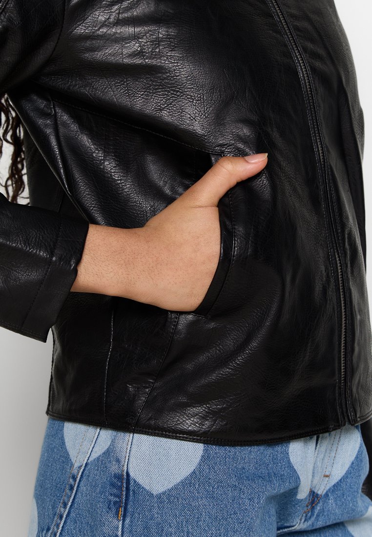 Chaqueta de cuero negra con cierre de cremallera, bolsillo parcialmente visible y acabado texturizado, combinada con jeans de mezclilla azul claro con estampado.