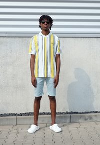 Polo a strisce gialle, bianche e nere; vestibilità rilassata. Pantaloni corti azzurri e scarpe slip-on bianche. In piedi su una superficie pavimentata.