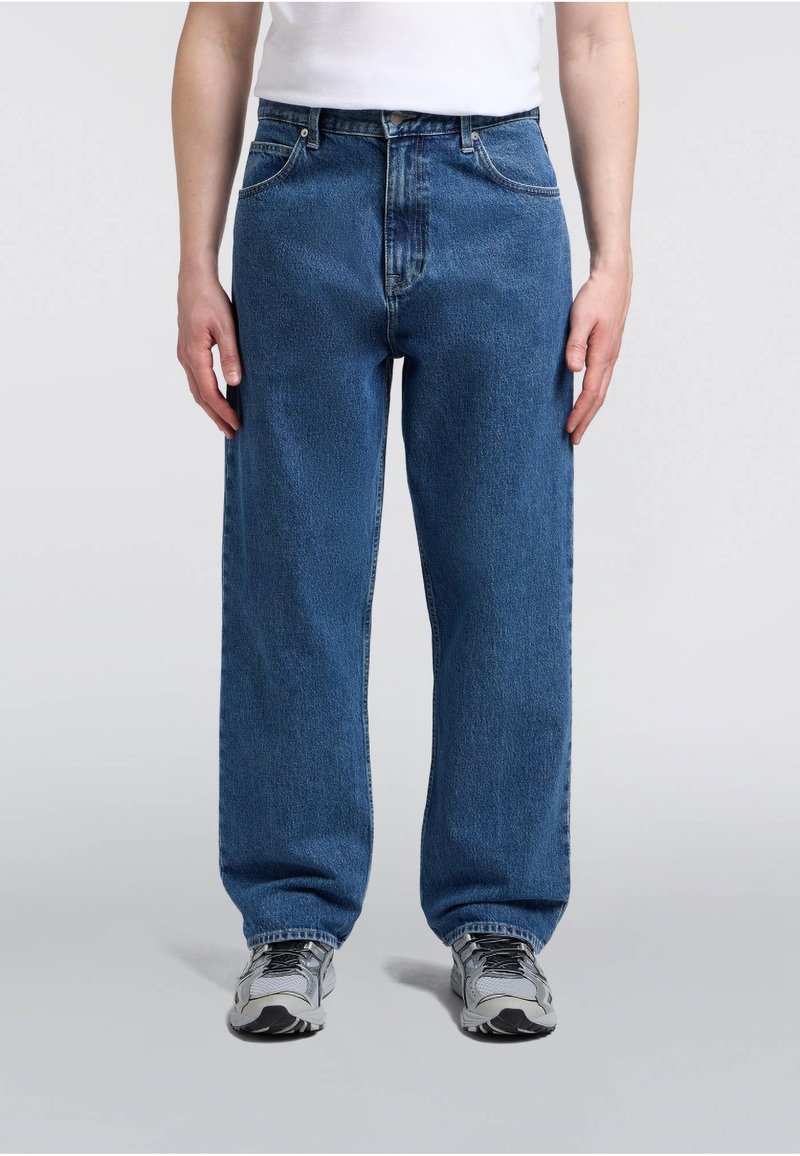 Personne portant un jean bleu droit, un haut blanc et des baskets grises, debout sur un fond uni gris clair.