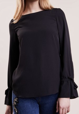 Kvinde iført en sort langærmet bluse med bindedetaljer ved ærmerne og blå jeans med dekorativ udsmykning på låret.