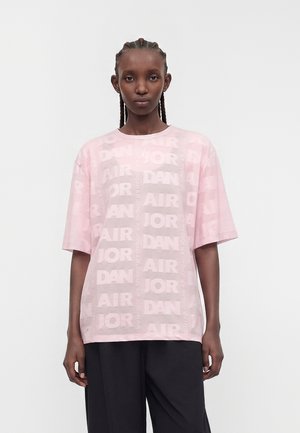 Sort kvinde med flettet hår iført en løs lyserød t-shirt med gentagende "Jordan Air" tekst og sorte bukser med vide ben, står mod en hvid baggrund.