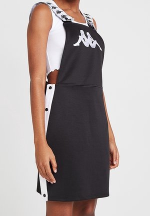 Nainen pukeutunut mustaan essumekkoon, jossa on valkoiset nappilistalliset sivupaneelit ja Kappa-logo edessä sekä olkaimet valkoisen hihattoman topin päällä.