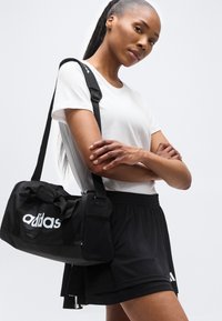 Γυναίκα που φοράει λευκό T-shirt και μαύρο σορτς με σταυρωμένα τα χέρια, κρατώντας μια μαύρη ταξιδιωτική τσάντα Adidas στον ώμο της, μπροστά σε έναν απλό φόντο.